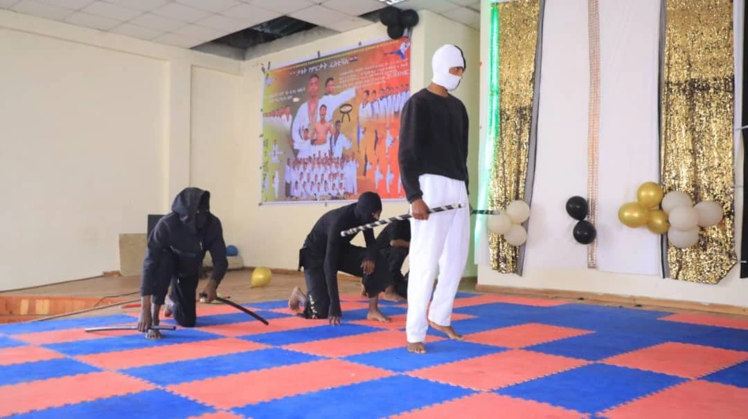 Participants kneeling on blue and coral mats beside a standing person in a decorated hall