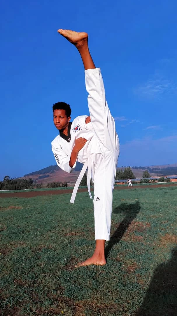Practitioner in a white Taekwondo uniform balancing in a high kick outdoors beneath a blue sky