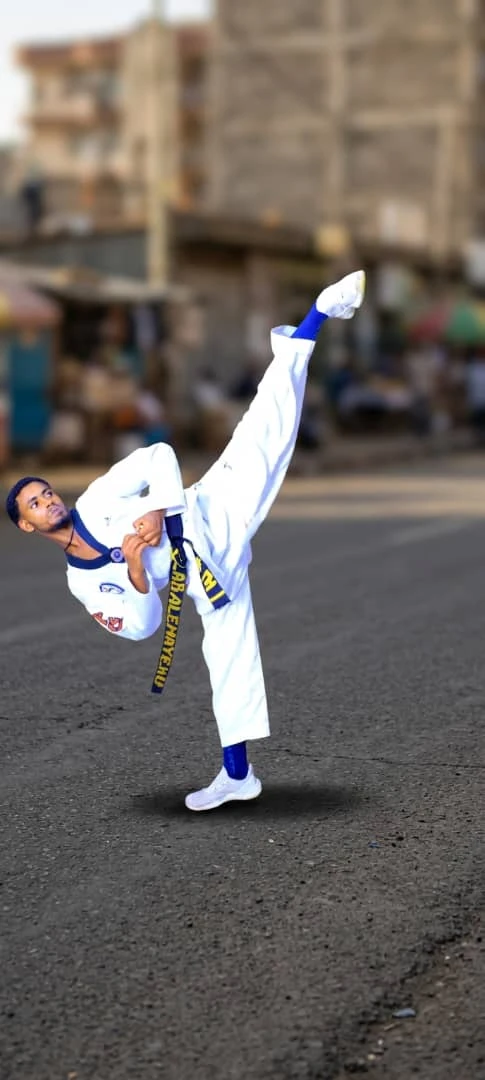 Taekwondo practitioner in a white uniform with blue accents performing a high kick on a street