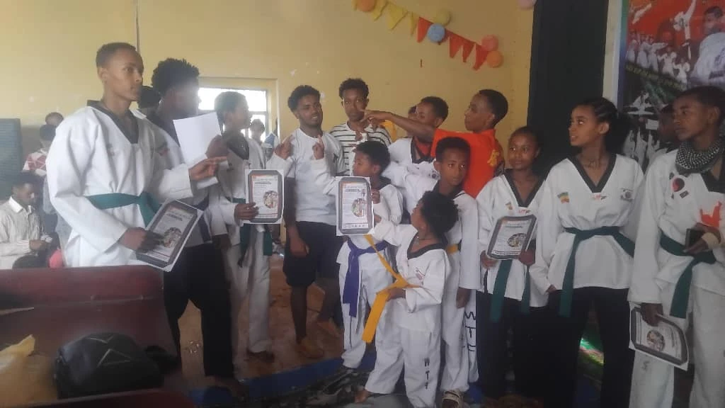 Group in white Taekwondo uniforms gathered indoors with several participants holding framed certificates