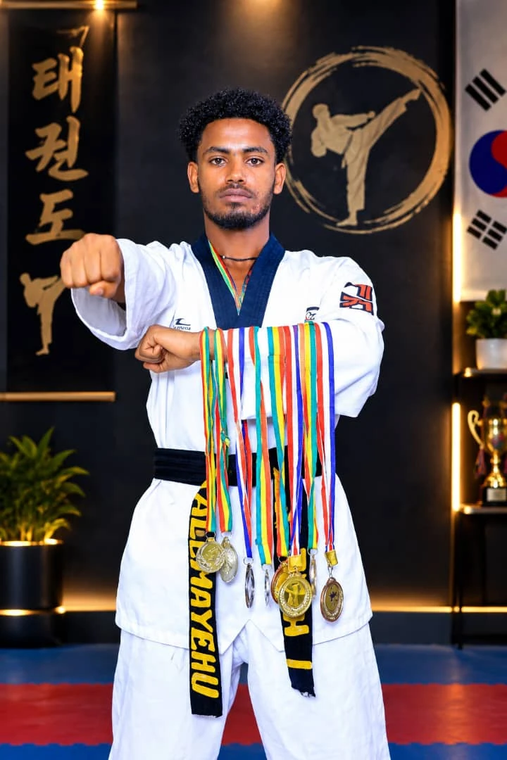 Taekwondo practitioner in a white uniform displaying medals with one fist extended before martial arts artwork