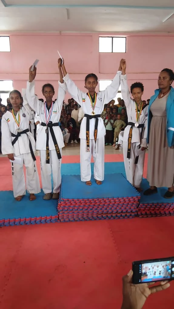 Four Taekwondo practitioners raising their arms on blue and red mats before seated spectators