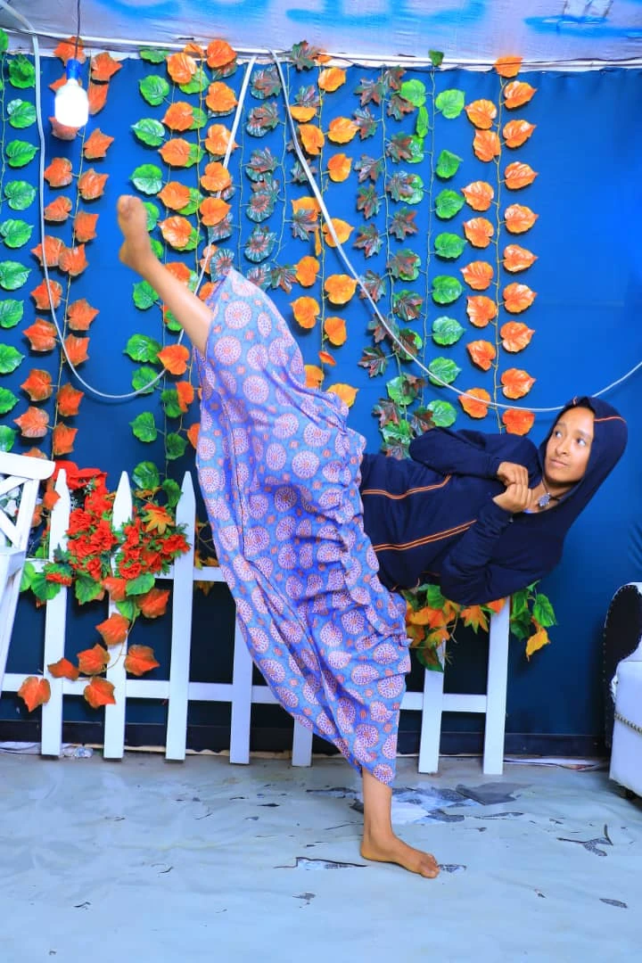 Person in a purple patterned dress balancing on one leg with the other raised beside another person against a blue leaf backdrop