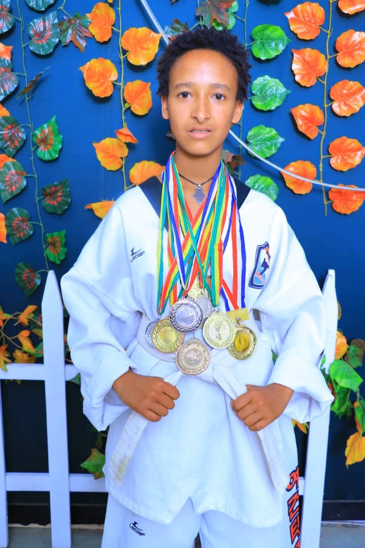 Martial artist in a white uniform wearing colorful medal ribbons before a blue backdrop with orange and green leaves