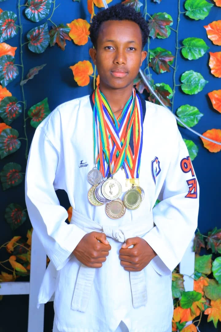 Person in a white martial arts uniform holding their belt while wearing medals before a vivid floral backdrop