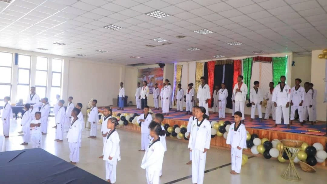 Large group of children and adults in white Taekwondo uniforms gathered in a hall with balloon garlands and a raised stage