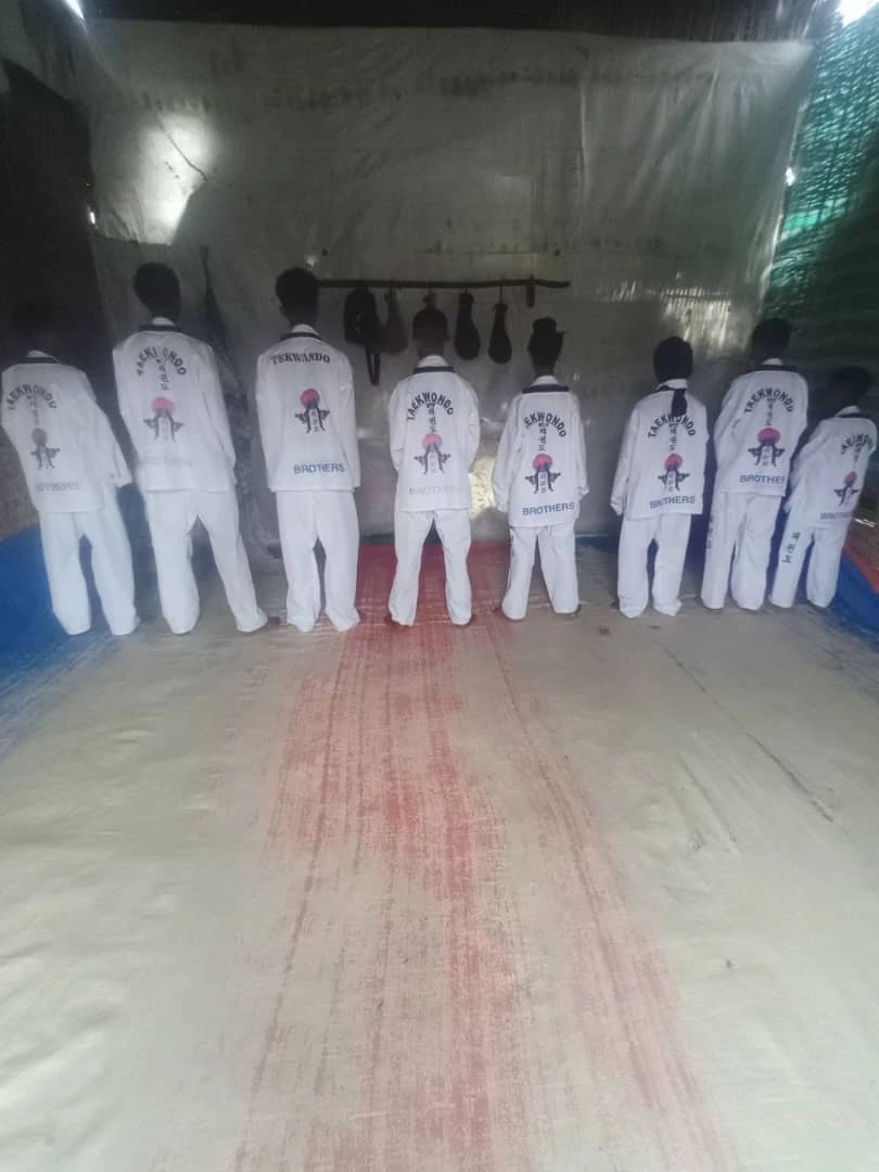 Taekwondo practitioners seen from behind facing hanging training bags inside a dojo with one participant kneeling